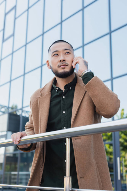 asiático hombre en otoño abrigo hablando en smartphone cerca de metal valla y edificio con fachada de vidrio - Foto, imagen
