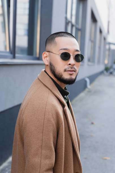 bearded asian man in autumn coat and sunglasses looking at camera near blurred building - Fotografie, Obrázek