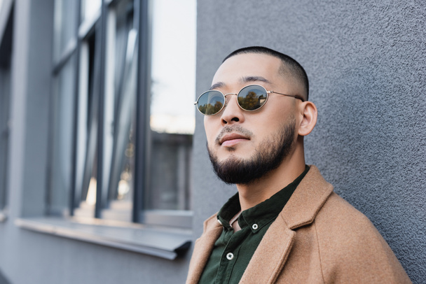 bearded asian man in autumn clothes and sunglasses standing near grey building  - Фото, зображення