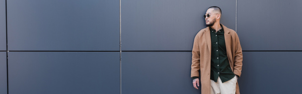 elegante uomo asiatico in cappotto beige e occhiali da sole in piedi con mano in tasca vicino alla parete grigia, banner - Foto, immagini