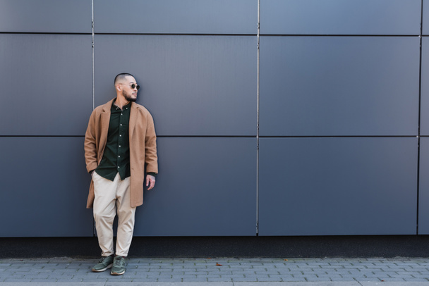 full length view of asian man in stylish autumn clothes standing with hand in pocket near grey wall - Photo, Image