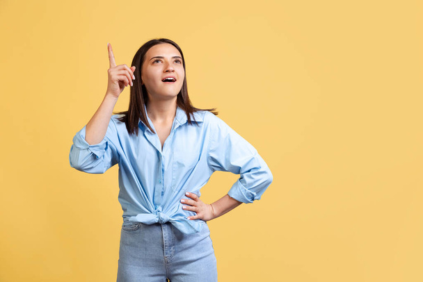 Cropped portrait of young beautiful woman in blue shirt pointing finger upwards isolated over yellow background. Ideas generation - Zdjęcie, obraz