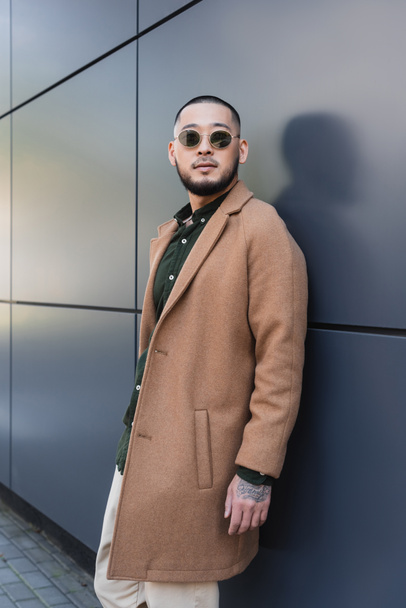 bearded asian man in trendy sunglasses and coat looking away near grey wall - Photo, Image