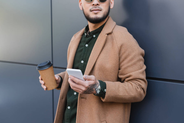 vista ritagliata dell'uomo in cappotto beige in piedi con caffè per andare e smartphone vicino alla parete grigia - Foto, immagini