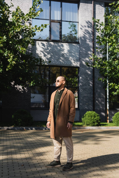 full length view of asian man in stylish autumn outfit standing near building and trees on street - Foto, afbeelding