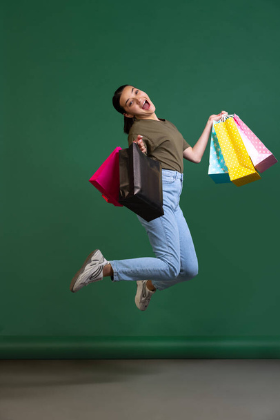 Full-length portrait of young beautiful happy woman in casual cloth jumping with shopping bags isolated over green background - Photo, image