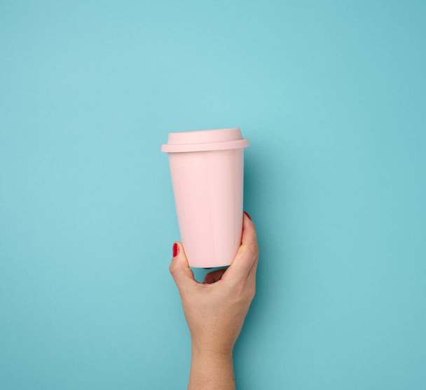 Mavi arka planda plastik kapaklı pembe bir seramik bardak tutan bir kadın el, mola zamanı ve kahve içmek - Fotoğraf, Görsel