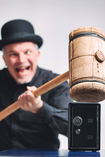 Gleeful senior man in bowler hat trying to smash a small metal safe or piggy bank with a large wooden mallet in a conceptual image with focus to the safe - Photo, Image