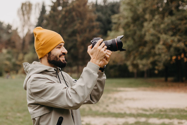 stylish bearded photographer with a camera in his hands takes a photo in the autumn park - Фото, зображення