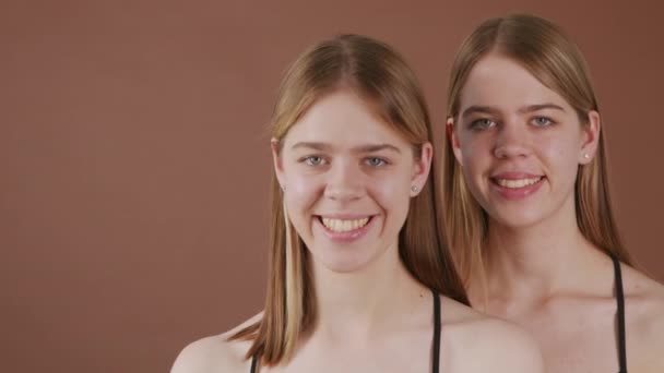 Schwenkporträtaufnahme glücklicher junger Zwillingsschwestern, die vor braunem Hintergrund in die Kamera lächeln - Filmmaterial, Video