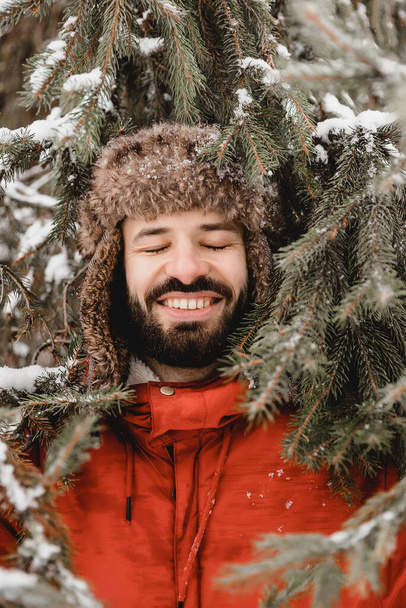 Stylish bearded man in a winter hat earflaps stands in a coniferous forest in frosty weather and smiles nicely. Frost winter season. Copy space - Zdjęcie, obraz