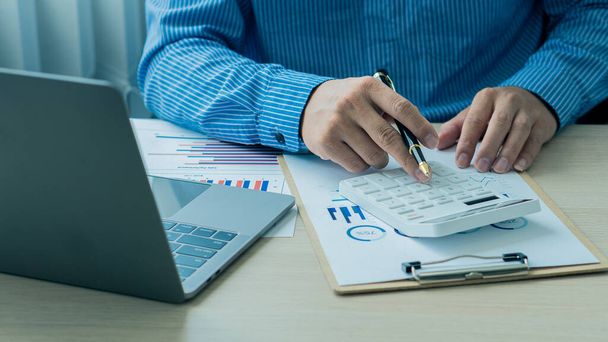 Een zakenman houdt een pen vast en drukt op een rekenmachine om maandelijkse bedrijfsrapporten of kantoorwinsten te berekenen en te analyseren met een laptop op zijn bureau. financieel bedrijfsboekhoudingsconcept - Foto, afbeelding