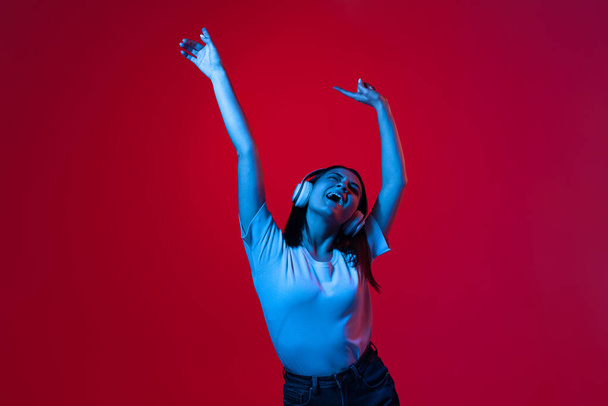 Retrato recortado de una joven hermosa mujer en camiseta blanca escuchando música en auriculares y bailando aislada sobre fondo rojo en luces de neón - Foto, imagen