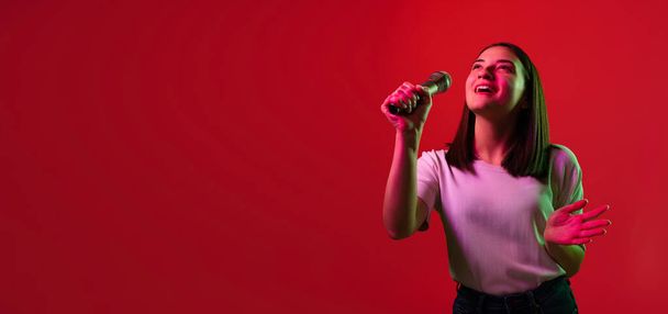 Cropped portrait of young beautiful woman in white T-shirt singing in microphone isolated over red background in neon lights. FLyer - 写真・画像