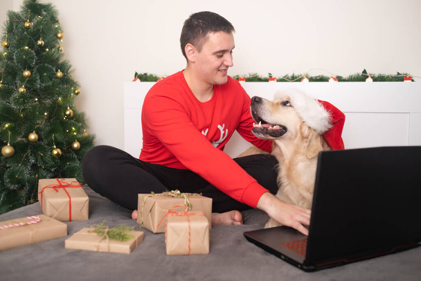 A man with a dog wearing a santa claus hat looks at a laptop screen. Christmas video greetings on social networks. New Year's home holiday. Happy New Year and Merry Christmas. - Foto, immagini