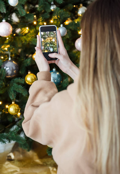 Teenage girl taking pictures of a Christmas tree on a smartphone. Christmas tree on the livingroom - Zdjęcie, obraz