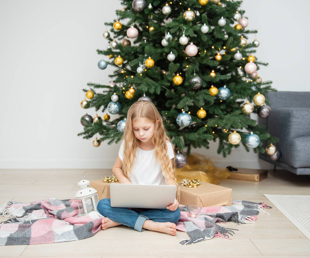 Weihnachtsbaum in einem großen Wohnzimmer. Kleines Mädchen spielt in der Nähe des Weihnachtsbaums. Ein Mädchen mit Laptop kommuniziert mit Freunden. - Foto, Bild