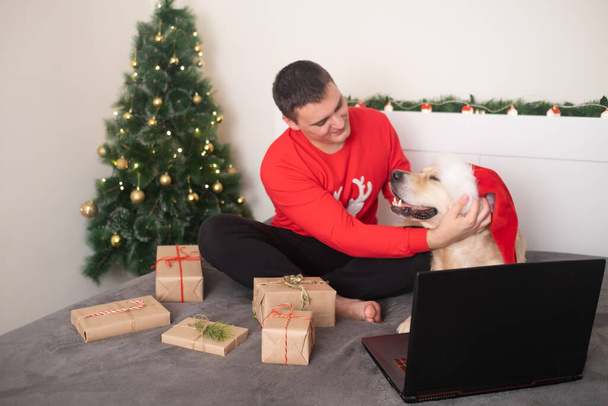 Un homme avec un chien portant un chapeau de Père Noël regarde un écran d'ordinateur portable. Salutations vidéo de Noël sur les réseaux sociaux. Vacances à la maison du Nouvel An. Bonne année et Joyeux Noël. - Photo, image