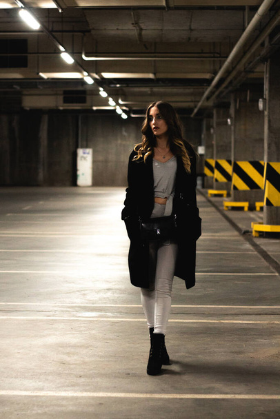 Atractiva joven morena posando en un aparcamiento subterráneo. Modelo de niña en abrigo negro posando en un aparcamiento subterráneo vacío. La foto a la moda de la muchacha joven hermosa con el maquillaje de tarde - Foto, Imagen