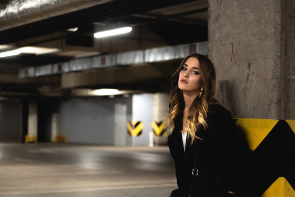 Attractive young brown-haired woman posing in an underground parking. Girl model in black coat posing in an empty underground parking. Fashionable photo of young beautiful girl with evening make-up - Fotografie, Obrázek