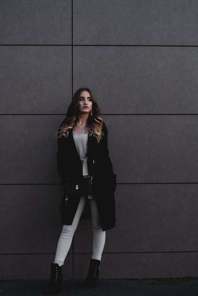 Sexy girl posing against a dark wall in the evening. An elegant fashionable girl in a long black coat, white jeans, with a leather bag stands near the wall of the building. Beautiful girl for a walk - Foto, Imagen