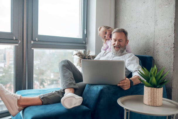 A mature man with a laptop sitting near his little cute daughter - Fotoğraf, Görsel