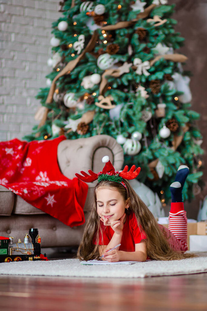 Funny cute caucasian girl left-handed writes a letter to Santa Claus near the Christmas tree. Happy childhood, time of miracles and desires. Merry Christmas. - Фото, изображение