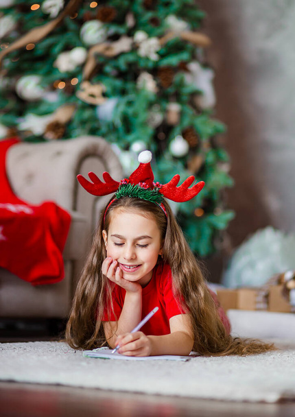 Funny cute caucasian girl left-handed writes a letter to Santa Claus near the Christmas tree. Happy childhood, time of miracles and desires. Merry Christmas. - Foto, Imagen