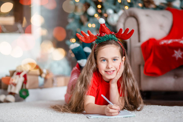 Funny cute caucasian girl writes a letter to Santa Claus near the Christmas tree. Happy childhood, time of miracles and desires. Merry Christmas. - Fotografie, Obrázek