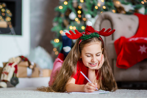 Funny cute caucasian girl writes a letter to Santa Claus near the Christmas tree. Happy childhood, time of miracles and desires. Merry Christmas. - Photo, image