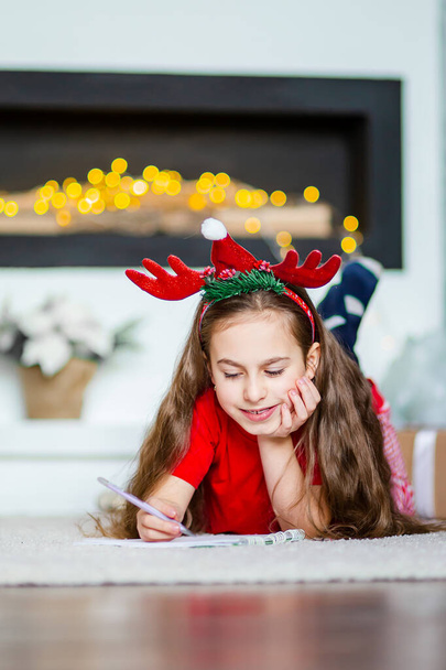 Funny cute caucasian girl writes a letter to Santa Claus near the Christmas tree. Happy childhood, time of miracles and desires. Merry Christmas. - Фото, изображение