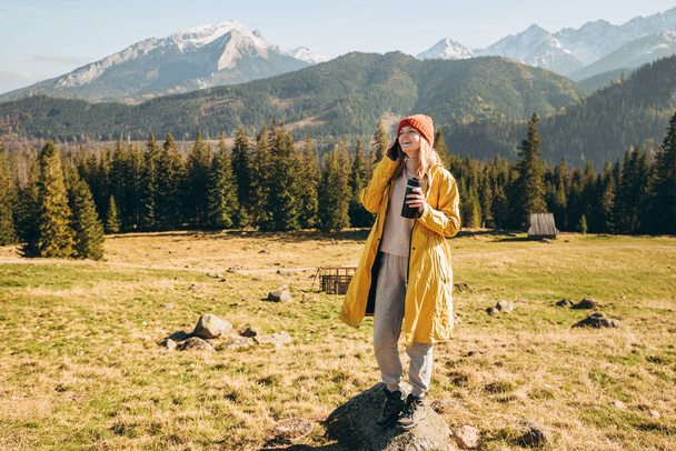 山の中でスマホで話題の魔法瓶カップを持つ若い金髪の女性。自由、幸福、旅行や休暇の概念、屋外活動。ハッピーガールは赤い帽子と黄色のジャケットを着て - 写真・画像