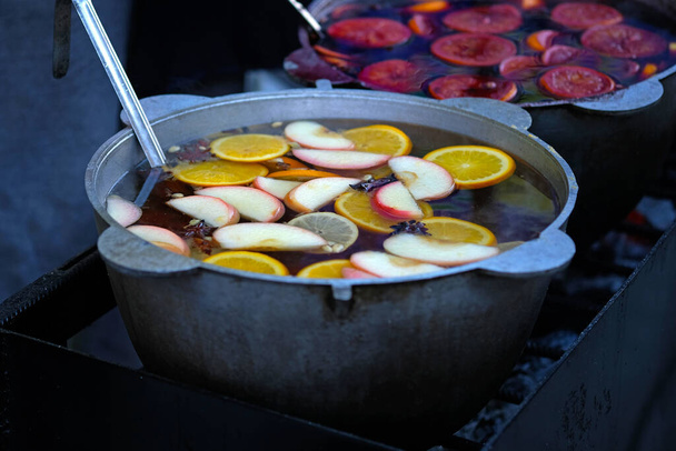 Large cauldron with mulled wine and fruit. Apples, spices and citrus fruits in a pan with mulled wine. Oranges and apples in wine close-up. Street food and drinks during the fair.  - Φωτογραφία, εικόνα