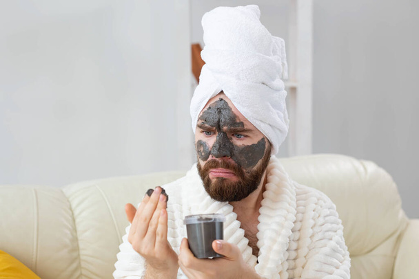 Brodaty, zabawny facet z maską kosmetyczną na twarzy zrobioną z czarnej gliny. Mężczyźni pielęgnacja skóry, humor i spa w domu koncepcja - Zdjęcie, obraz