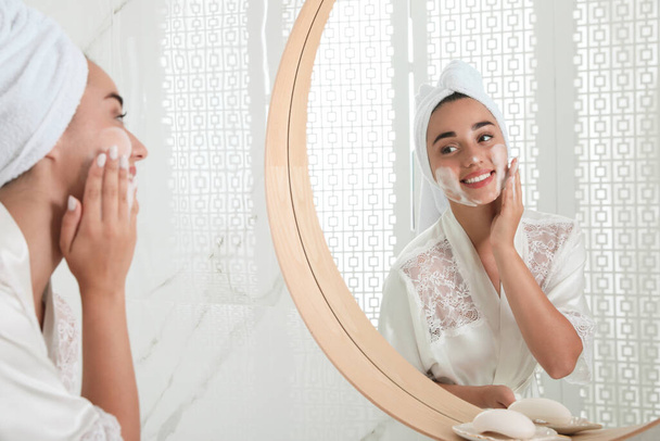 Beautiful young woman applying cleansing foam onto face near mirror in bathroom. Skin care cosmetic - Φωτογραφία, εικόνα