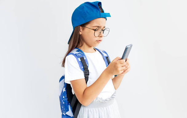 Cute kid wearing blue cap, white t-shirt, transparent eyeglasses, and backpack holds smartphone on a white background. Little girl playing online games on mobilephone. Kid learning online. - Foto, Imagem