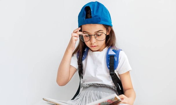 Όμορφο κοριτσάκι με μπλε καπέλο και λευκό t-shirt με σακίδιο κάθεται στο πάτωμα και διαβάζοντας ένα βιβλίο. Μαθήτρια με διάφανα γυαλιά που μαθαίνει από το βιβλίο. - Φωτογραφία, εικόνα