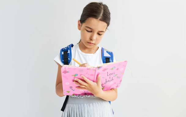 Studio portret uczennicy z niebieskim plecakiem pisze coś na jej różowym notatniku podczas pozowania na białym tle studio. - Zdjęcie, obraz