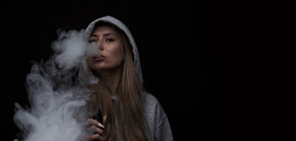 Portrait of beautiful blonde girl in gray hoodie smokes vape isolated on black studio background, cloud of steam smoke, mini hookah - Fotó, kép
