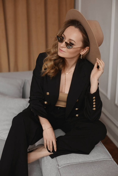 A beautiful model woman in a black suit, wide-brimmed beige hat and trendy sunglasses poses on the sofa - Foto, Imagen