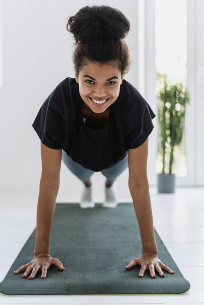 Pionowy widok szczęśliwej afro amerykańskiej kobiety patrzącej w kamerę, uśmiechającej się szeroko, wykonującej codzienny trening sportowy, podnoszącej się z podłogi. Silna dziewczyna stojąca na desce, ćwicząca w domu - Zdjęcie, obraz
