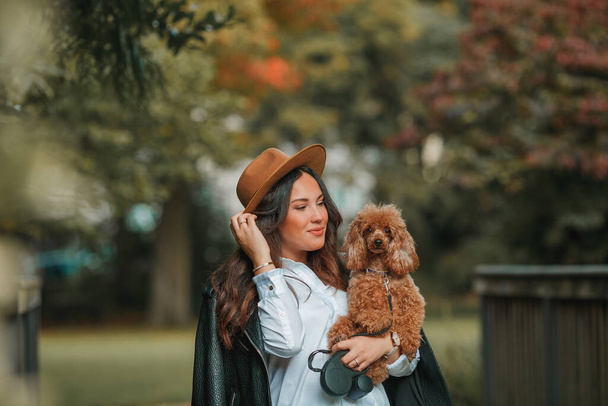 Uma bela jovem mulher de cabelos escuros caminha no parque e abraça seu cachorro poodle. Humor de outono. Queda. - Foto, Imagem
