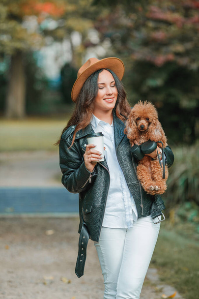 美しい若い暗い髪の女性が公園を歩き、彼女のプードル犬を抱きかかえています。秋の気分。秋. - 写真・画像