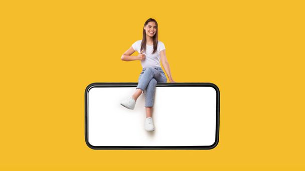 Femme assise sur l'écran blanc vide du smartphone montrant pouces vers le haut - Photo, image