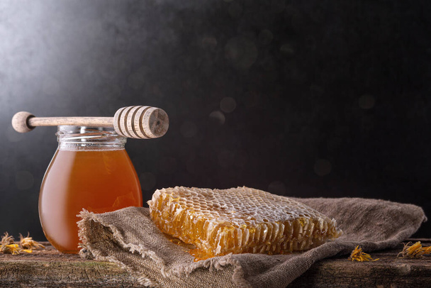 Honey background. Sweet honey in the comb, glass jar and a spoon for honey on the table. Dark background. - Фото, изображение