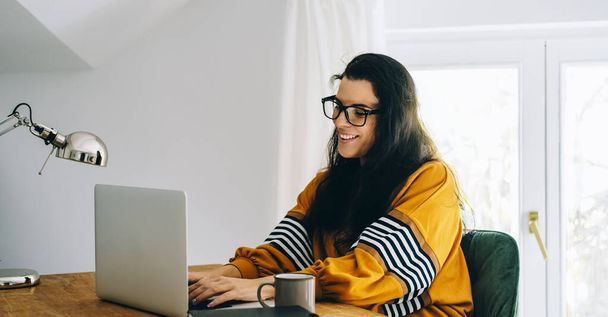 Μια χαμογελαστή γυναίκα στο γραφείο της έχει βιντεοκλήση στον υπολογιστή. Γυναίκα ανεξάρτητη υπάλληλος και εργασία εξ αποστάσεως μορφή σπίτι. Μαμά blogger χρησιμοποιώντας internet shop. - Φωτογραφία, εικόνα