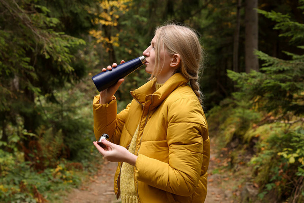 Young woman drinking hot beverage from thermo bottle in autumn forest - Photo, image