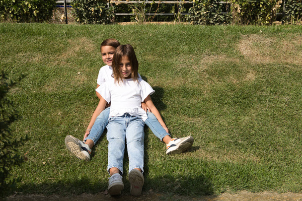Portrait de deux petits frères et sœurs enfants couchés sur l'herbe. Ils sont vêtus de T-shirts et de jeans blancs. La sœur aînée est une fille transsexuelle. Concept d'une famille unie et heureuse. - Photo, image