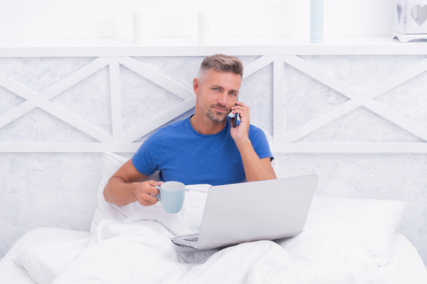 Happy man talk on cellphone drinking coffee and using laptop in bed in morning, mobile - Foto, Imagem