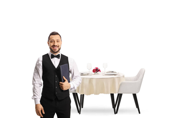 Waiter with a bow tie holding a menu list in front of a restaurant table isolated on white background - Photo, Image